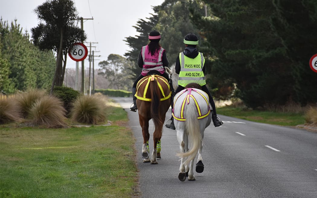 Two horseback riders riding down a paved country road wearing hi-vis vests wit 'PASS SLOW AND WIDE' written on the back.  There is a sixty kilometre per hour sign in the background.