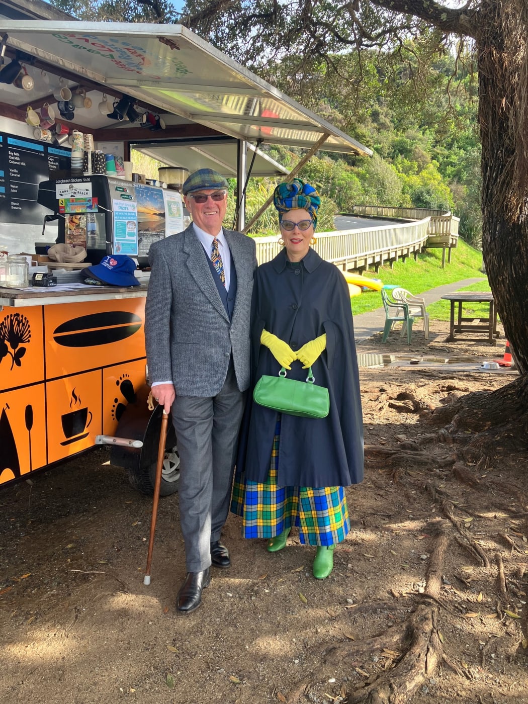 Jacqui and her husband stand outside a food truck in their vintage clothes. Jacqui wears a navy cape, blue and yellow tartan skirt, yellow gloves, a green bag, and a matching tartan bonnet.