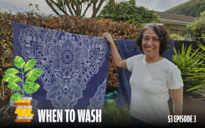A cheerful woman in a white T-shirt stands outside next to a clothesline, holding a large, dark blue towel with a detailed white floral pattern. Greenery and trees are in the background, creating a natural setting. In the lower left, a graphic of a potted plant growing from a stack of coins is displayed, along with the text 'WHEN TO WASH' in bold white letters and 'S3 EPISODE 3' in smaller white text on the right.