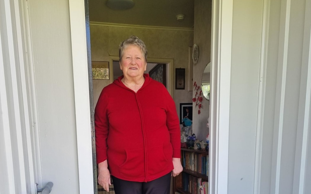 Elizabeth Gedye standing in her doorway. She wears a bright red top, black pants and striped socks.