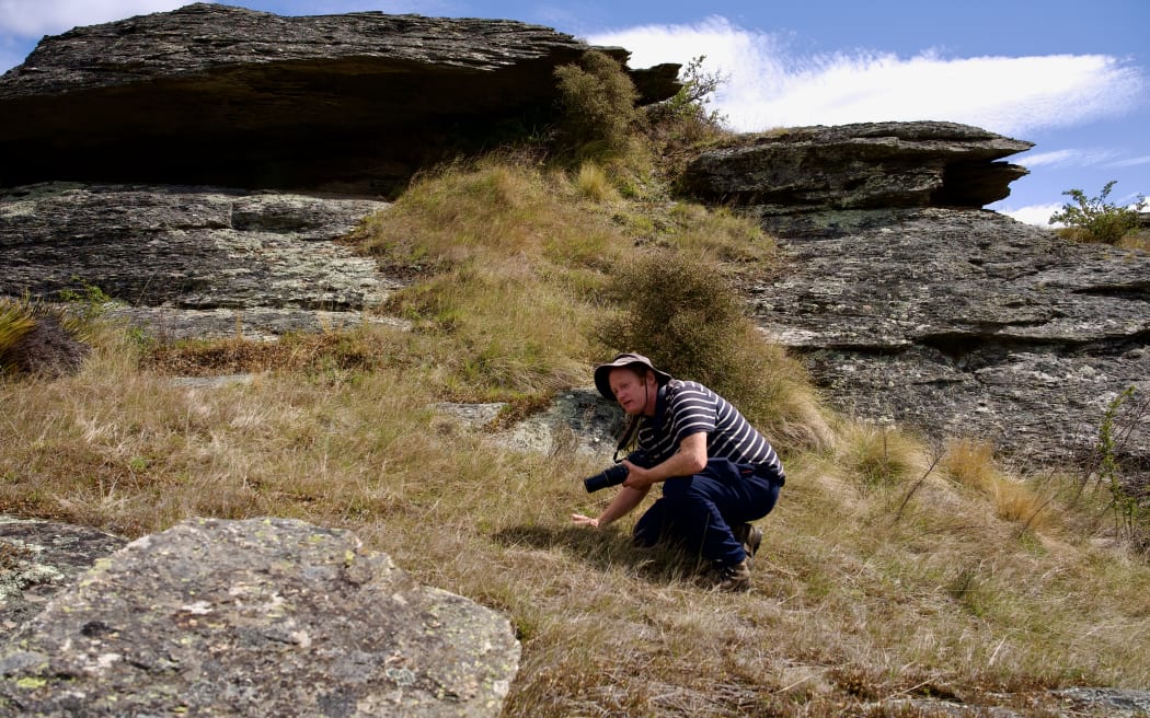 A man in a stripey t-shirt and wide-brimmed hat crouches in dry brown grass in front of a large flat rock formation.