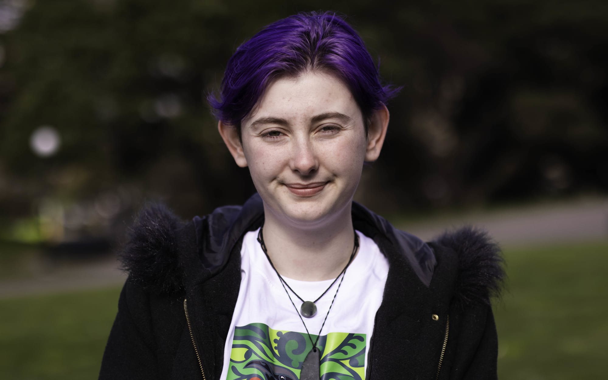 Maxine, a young woman with short purple hair and two pieces of pounamu around her neck, stands outside in front of foliage which is blurred in the background.