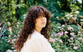 Lilly Carron has long flowing curley brown hair. She is wearing a white gown and standing in front of a background that looks a bit like an english flower garden.