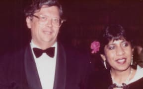 David and Jeya stand side by side, smiling. They are both in formalwear, David in a black suit jacket and black bowtie, and Jeya in a pink dress with a black cape over the top.