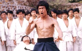 Bruce shirtless, mouth, in pose, tournament fight, on Han's Island, action shot in ETD. Students, extras, flags, stone wall in background. Iconic portrait, punch behind.