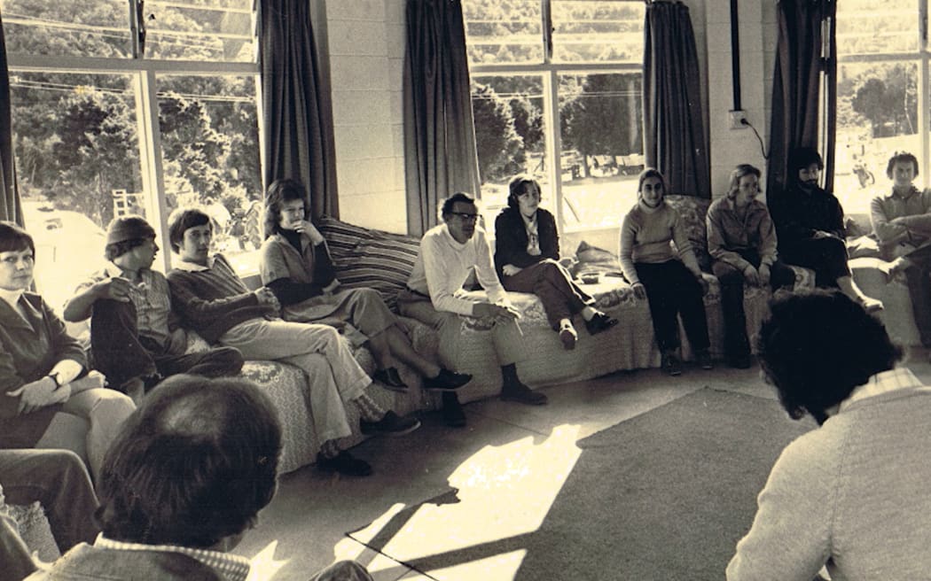 An old photo. A room full of people sitting on couches around the perimeter of the room, listening to someone speak.