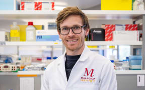 A headshot of Tom, he is wearing glasses and a white labcoat with a Malaghan Institute logo. He is standing in a lab in front of several shelves stocked with lab equipment and bottles.