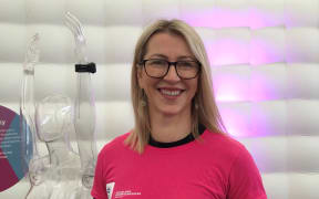 Portrait shot of Professor Tawhai. She wears a pink ABI t-shirt, with her hair down and glasses. She is smiling. Behind here is a white divider background and a transparent human model.