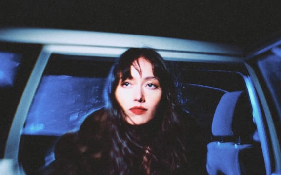 Fazerdaze - a young woman with long dark hair and lipstick - looks out from an open car door.