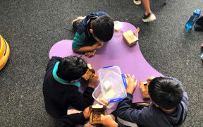 A group of children sit around a low, purple table eating school lunches out of brown cardpaper boxes.