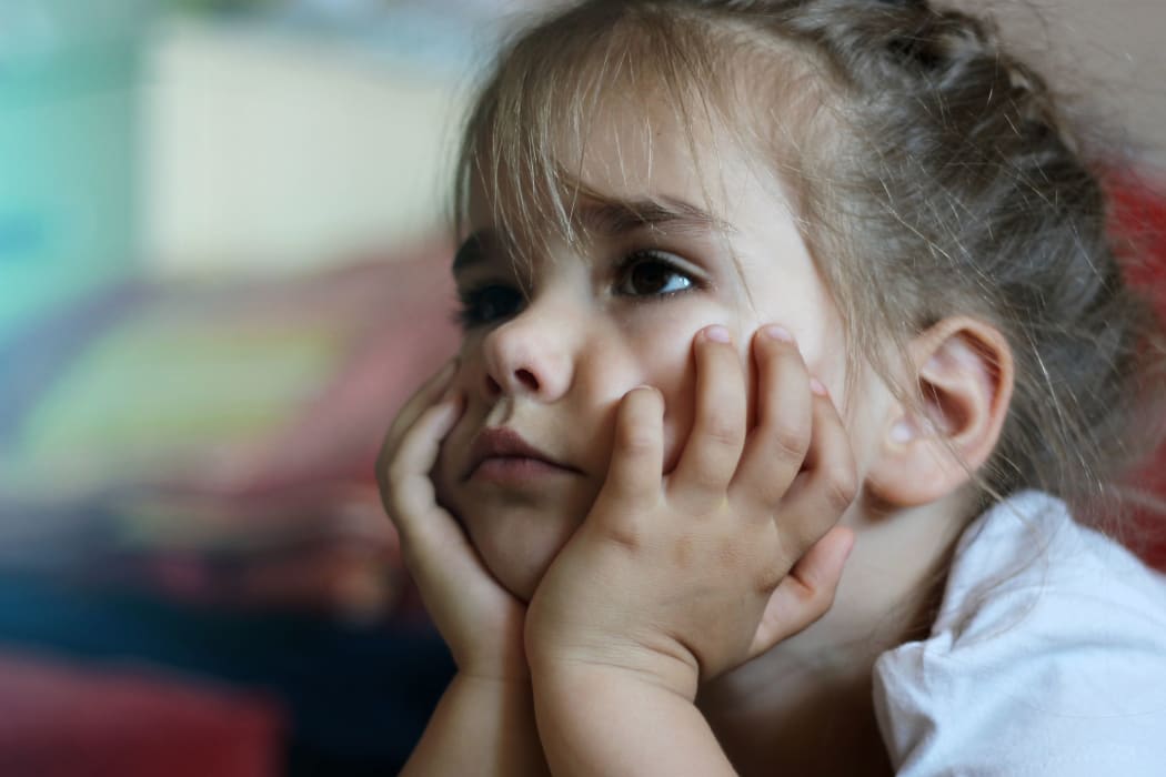 A photo of a little girl lying on the sofa with hands on chin watching the television and looking worried