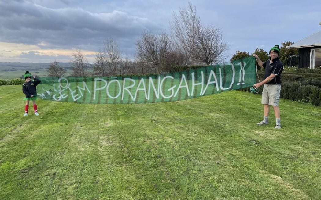 A man standing on a grassy field holds a sign saying Go get em Porangahau!