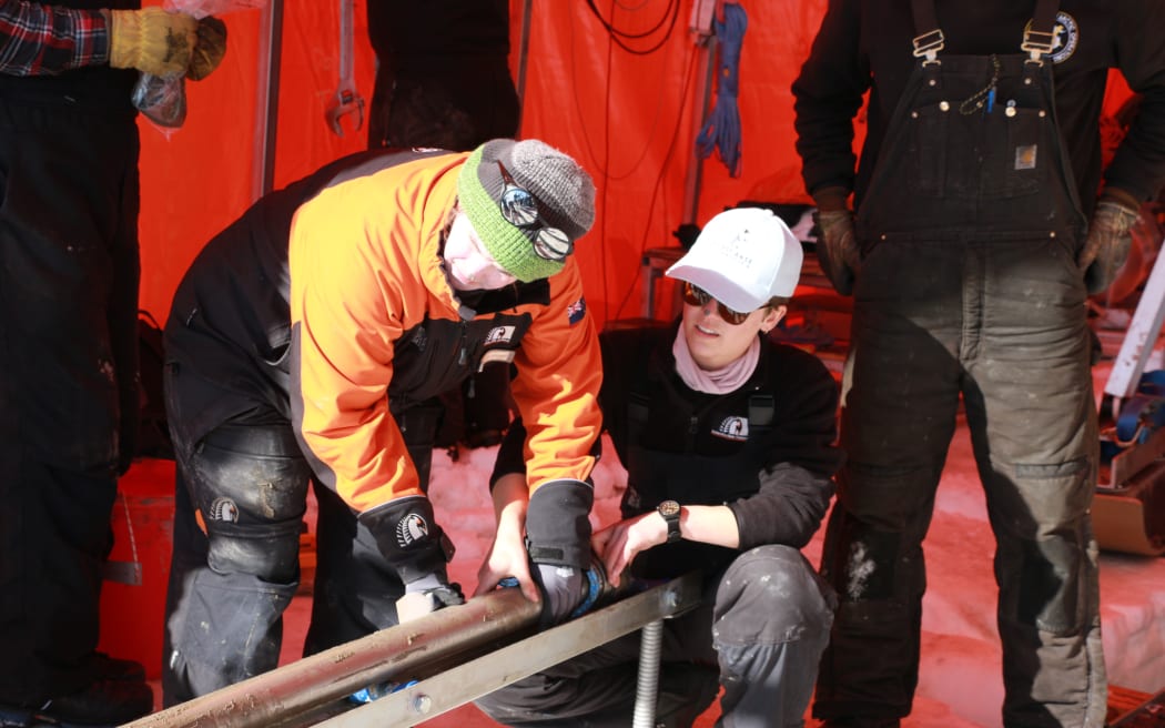 Two people hold a long cylinder in a metal gutter. One is standing and bending over, wearing a beanie and an orange and black jacket. The other is kneeling and wearing sunglasses and a white cap. They are standing in an orange tent.