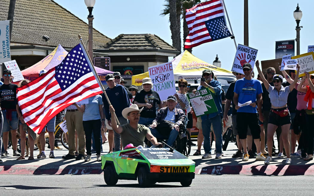 A person waving a US flag drives a toy Tesla Cybertruck past demonstrators gathered for a 'Hands Off!' protest against the policies of US President Donald Trump and his advisor, Tesla CEO Elon Musk, in Huntington Beach, California.