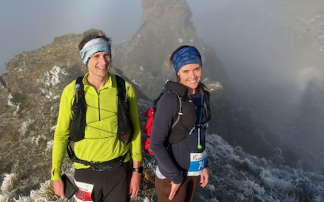 Two people in outdoors gear wearing backpacks are smiling, standing atop a craggy mountain peak shrouded in mist.