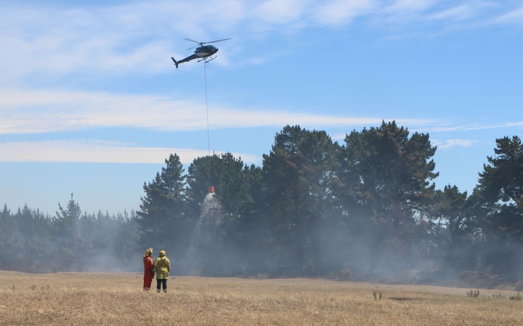 Firefighters watch on as a helicopter dumps water on a wildfire near Kirwee, Canterbury. February 5, 2024.