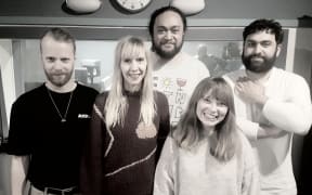 Five band members standing in the RNZ recording studio against the control room window