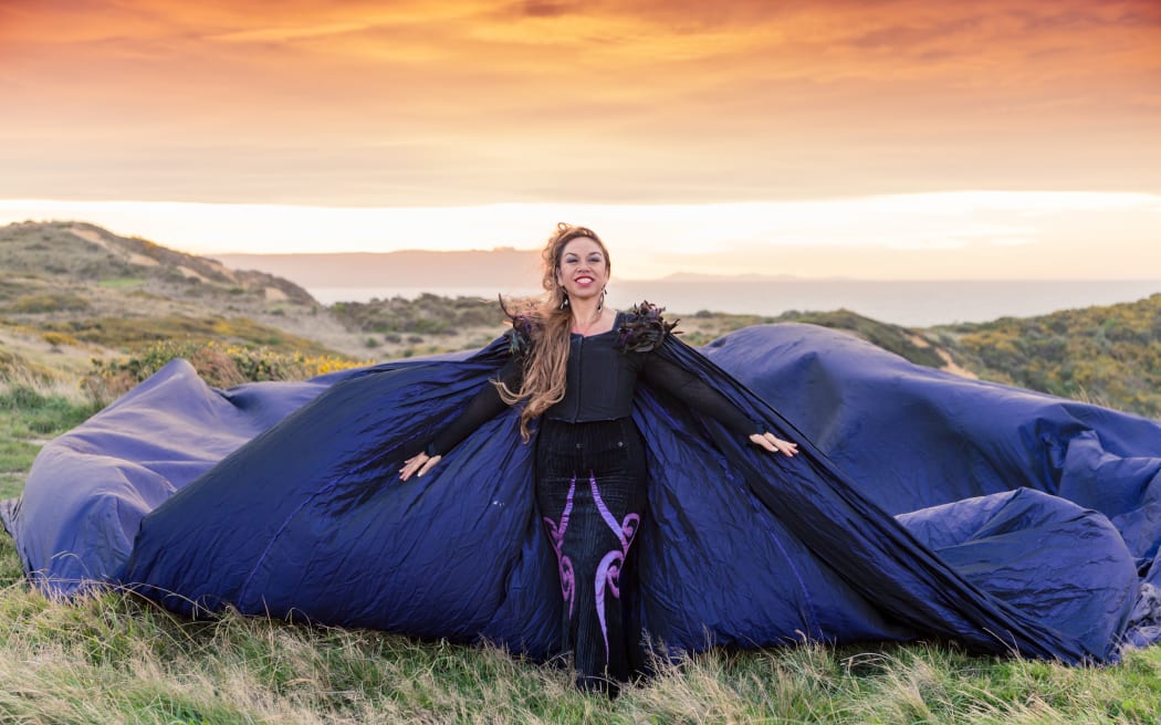 Toni Huata stands on top of a mountain wearing a purple cape that is billowing in the wind.