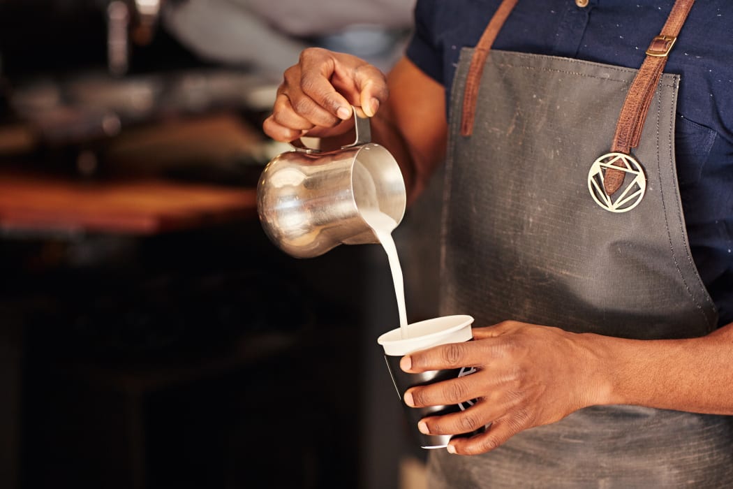 Cropped image of an African barista carefully pouring milk from a stainless steel jug into a takeaway cup in a coffee shop