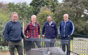 Four men stand behind a wire cage trap.