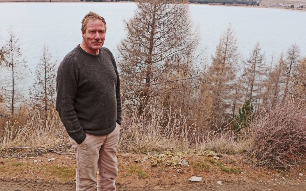 Man in jersey stands on farm land with lake in background.