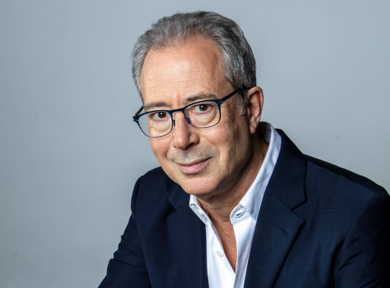 A head and shoulders shot of comedian Ben Elton, who has grey hair, glasses and is wearing a white collared shirt with a navy jacket.