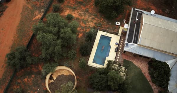 An aerial shot of a person swimming in a small pool with buildings and red dirt around it.
