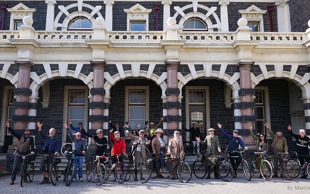 A group of people pose on their bikes in front of the Dunedin Railway Station. Many are dressed in tweed.
