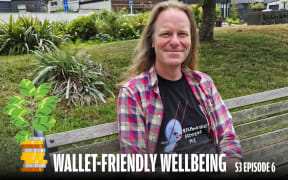 A man with long, light brown hair sits on a wooden bench outdoors, smiling at the camera. He wears a plaid shirt over a black T-shirt that reads 'Responsibility Saddens Me' with an illustrated character. The background features lush greenery, including bushes and grass, with a building partially visible. A money plant graphic is in the lower-left corner, accompanied by the text 'Wallet-Friendly Wellbeing S3 Episode 6.