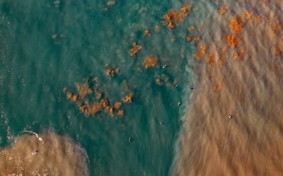 A drone shot above coastal water showing clear blue water on one side and brown, sediment-laden water to the right