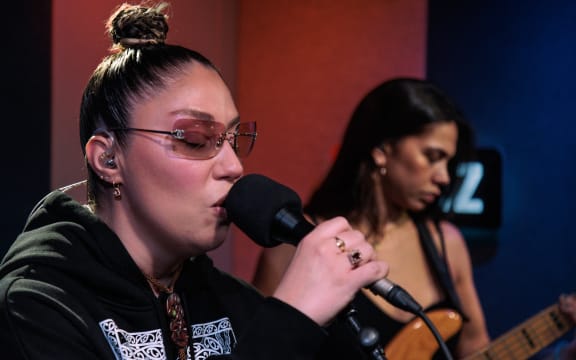 Theia is singing into a microphone. There is a bass player in soft focus behind her. In the background you can see the Z of some RNZ branding.