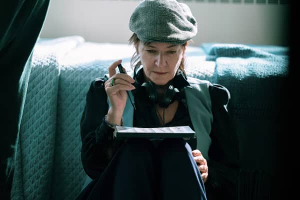 Lynne Ramsay in a grey hat and with headphones around her neck holds a pen and looks downward.