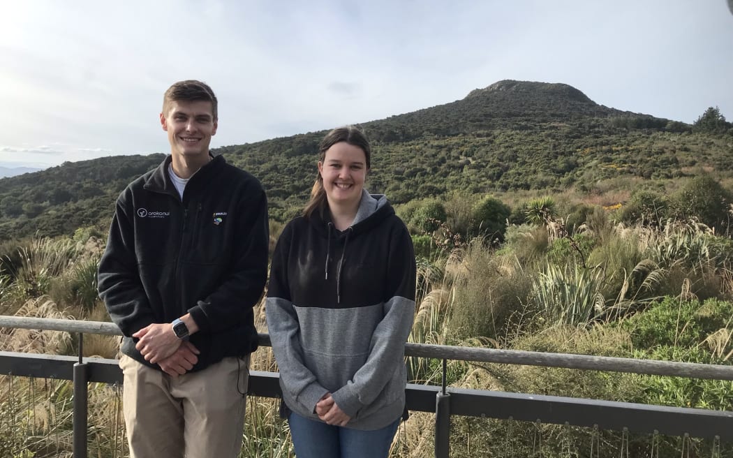 Taylor and Greer are standing on the verandah of Orokonui, smiling, with a bush-covered hill behind them.