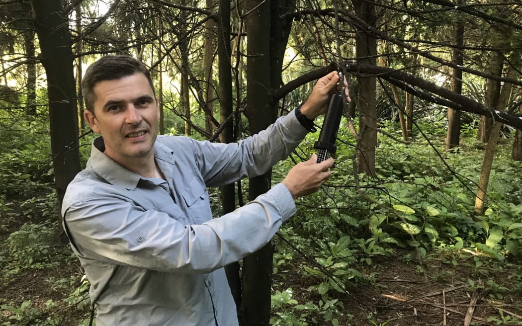 A man stands next to a tree pointing out a black plastic acoustic recording device.