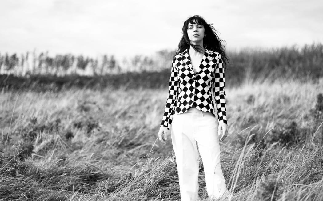 This is a black and white photo of Susan O'Neill. She is standing in a field of long grass. Susan wears a check shirt. It is impossible to say which colour because the photo is black and white. She has long, slightly wild hair. Judging by her hair it is moderately windy.