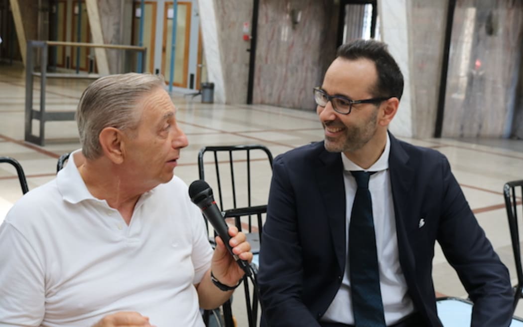 Loli and Francesco sit in two chairs in conversation together. Loli, wearing a white polo shirt, holds a microphone and is mid-speech. Francesco smiles, listening, wearing a suit.