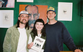 Sam, Rosa and Flynn pose for the camera. They are all smiling. Rosa holds a copy of Newzician magazine.