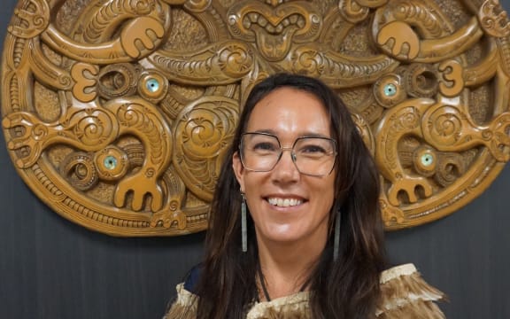Claire Charters is wearing glasses with long brown hair, she is standing up and smiling at the camera.