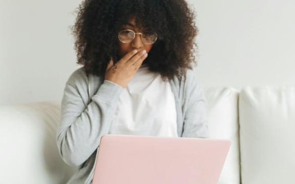 A woman wearing a white tshirt and grey cardigan covers her mouth with her hand in surprise as she looks at a laptop.ggen