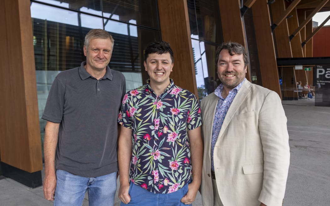 A head and torso portrait shot of the three men in front of a glass and wood framed building. Jacob is wearing a grey polo shirt and blue jeans, Zac is in a colourful flower shirt and jeans, and Vincent wears a cream suit with a blue shirt. All are smiling