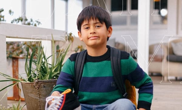 Elijah Tamati - a boy in a striped sweater and backpack - sits on some steps.