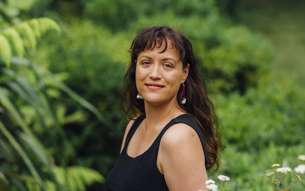 Māori architect Jade Kake wears a black tank top and pants and stands in front of greenery outside.