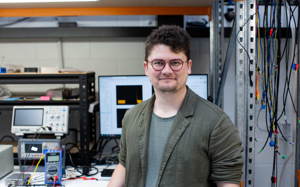 A man wearing glasses and a light blazer with the sleeves rolled up stands in a science lab in front of scientific equipment, cables and monitor screens.