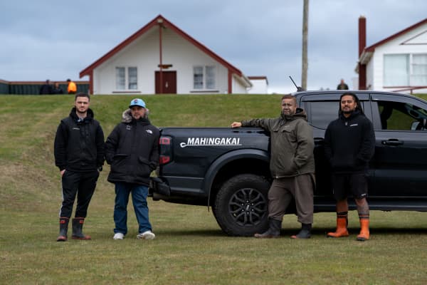 Left to right: Te Aorere Pewhairangi, Dr Rangi Mātāmua, Che Wilson, and Parekaia Tapiata, stand in front of a wharenui and ute which says 'Mahinga kai' on it.