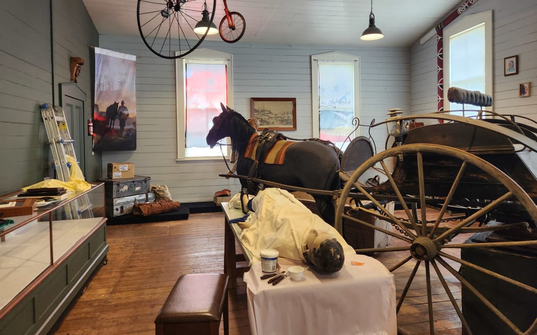 Interior of the Eketāhuna Museum, showing a mannequin lying on a table and life-size model of a horse with cart and a penny farthing hanging from the ceiling