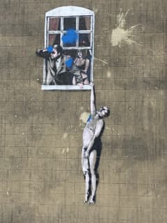 A painting on a wall depicts a naked man hanging out of a window which two people appear in.