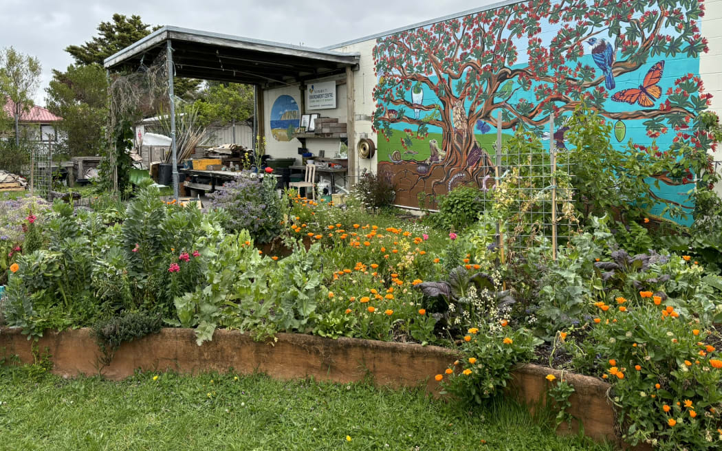 A building with a huge colourful mural of a tree and creatures painted on its wall, next to a verandah covering an array of tables and work benches. In the foreground, there is a veggie and flower garden bed bursting with life.