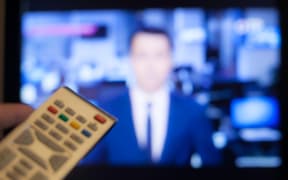 Hand holding TV remote control with a television in the background. Close up.