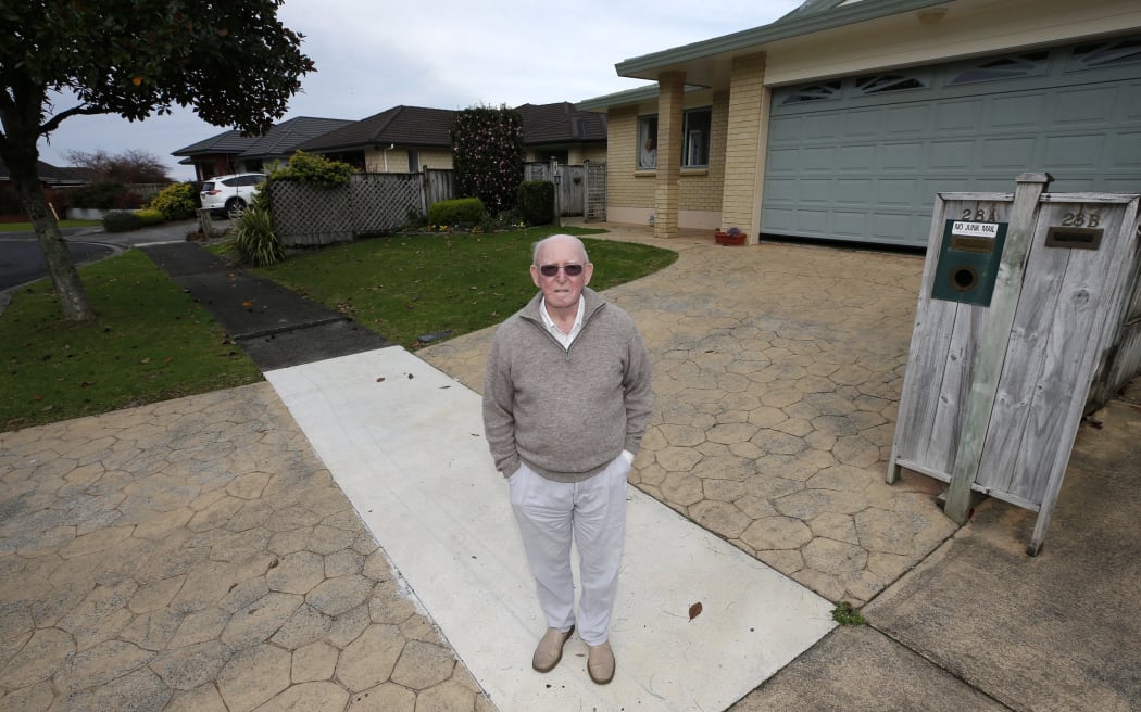 Roy Halford has had to pay $2000 to fix his driveway where it crossed council footpath damaged by the magnolia tree behind him. Temporary council footpath repairs can also be seen. He collects magnolia leaves several times a week, filling black plastic rubbish bags.