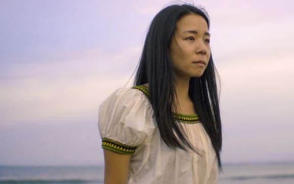 woman with long dark hair in a white top with sunset and ocean behind her
